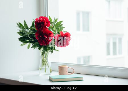 Un bellissimo bouquet di vivaci peonie rosse in un vaso di vetro, posto su un davanzale. La luce naturale soffusa esalta l'atmosfera accogliente della casa Foto Stock