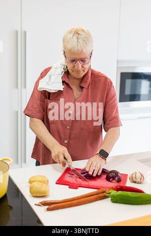 Una donna anziana prepara un pasto nella sua cucina moderna. Sta tritando verdure su un tagliere rosso, circondato da ingredienti freschi, abbracciando Foto Stock