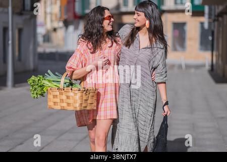 Due donne camminano braccio al braccio, sorridendo in abiti estivi. Uno porta un cestino di vimini pieno di verdure fresche, che indica una giornata di shopping allegra, CEL Foto Stock