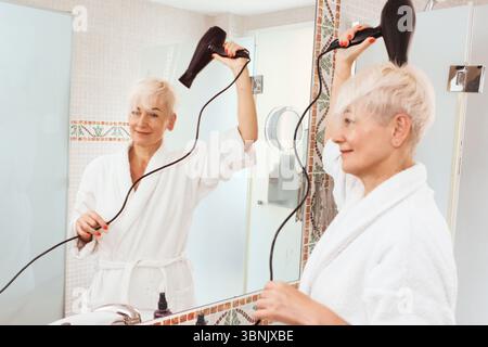 Una donna matura con capelli biondi corti utilizza un asciugacapelli mentre indossa un accappatoio bianco, riflesso nello specchio del bagno. L'ambiente trasmette un'atmosfera rilassata Foto Stock