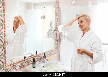 Una donna matura con i capelli biondi corti in un abito bianco sorride mentre si guarda nello specchio del bagno, creando un senso di routine mattutina e se stessa Foto Stock