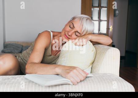 Una donna matura e serena con i capelli corti si gode un momento di riposo sul divano di casa. I suoi occhi sono chiusi mentre si appoggia su un cuscino morbido, creando un Foto Stock