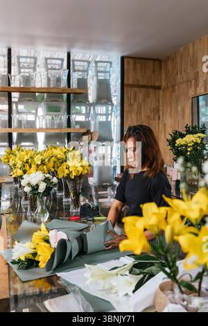 Un fiorista dedicato a Bali organizza con cura vivaci gigli gigli gialli e rose bianche in un meraviglioso bouquet. La luce del sole si riflette sui vasi di vetro, c Foto Stock