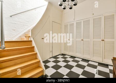 Un interno moderno caratterizzato da una scala in legno con un'elegante ringhiera in metallo che conduce ad un piano superiore. Il pavimento bianco e nero a scacchi aggiunge un tocco di stile Foto Stock