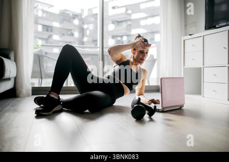 Una donna in abbigliamento sportivo si allena a casa su un pavimento di legno, utilizzando un computer portatile come guida. È impegnata in una routine di allenamento con un set di manubri Foto Stock