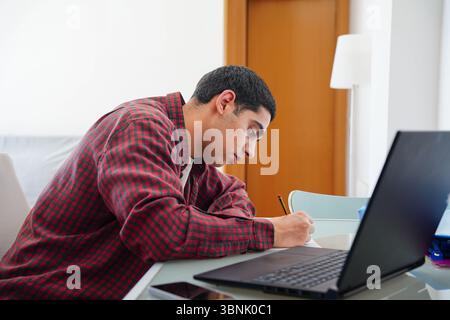 Un giovane arabo siede a un tavolo, profondamente impegnato a scrivere appunti accanto al suo portatile. L'atmosfera serena cattura un assaggio della sua routine quotidiana Foto Stock