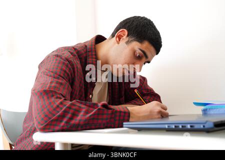 Un giovane arabo scrive intensamente in un quaderno, mostrando la sua dedizione agli studi. Seduto a una scrivania con un portatile, incarna la concentrazione e la concentrazione Foto Stock