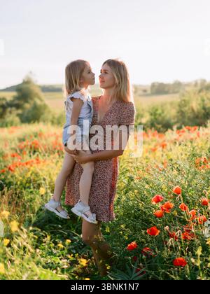 Madre felice che tiene la figlia in un campo di papavero in fiore in una giornata estiva di sole, godendo la natura e l'amore per la famiglia Foto Stock