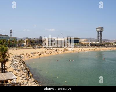 Una vivace scena sulla spiaggia di Barceloneta che guarda verso la città, con la torre Torre Sant Sebastià e una gru da costruzione, che indica la continua d Foto Stock