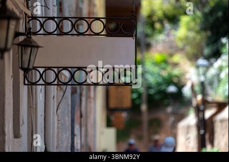 Un cartello bianco è appeso su un muro in una strada tranquilla con luce solare e vegetazione sparse. Foto Stock
