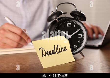 Nota cartacea con scadenza della parola sulla sveglia e uomo che lavora al tavolo di legno, messa a fuoco selettiva Foto Stock