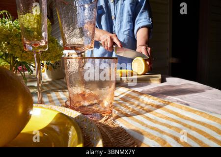 Primo piano di una donna che affetta un limone su una tavola di legno. La luce del sole mette in risalto agrumi freschi, bicchieri e un'accogliente atmosfera estiva, perfetta per limonata o b b Foto Stock