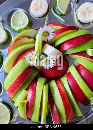 Frutta fresca su piatti da portata, nutrizione ricca di vitamine, banane sane affettate, limes e mele con buccia rossa e verde come antipasti, margherite come decorazione Foto Stock