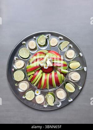 Frutta fresca su piatti da portata, nutrizione ricca di vitamine, banane sane affettate, limes e mele con buccia rossa e verde come antipasti, margherite come decorazione Foto Stock