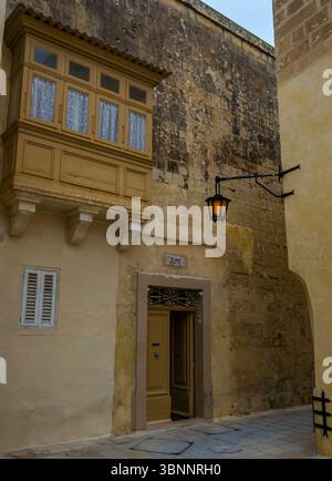 Mdina, Malta - 10 febbraio 2024: Casa medievale in pietra calcarea con balcone in legno e porta aperta illuminata con lanterna in una giornata di pioggia cupa in inverno Foto Stock