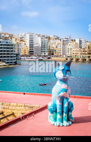 Sliema, Malta: 11 febbraio 2024: Gigante statua blu del gatto seduta su un tetto rosso nei Giardini dell'indipendenza con la baia di San Giuliano sullo sfondo Foto Stock
