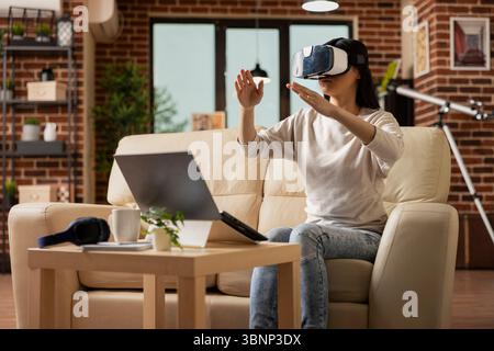 Donna d'affari seduta sul divano, indossando un visore vr e interagendo con l'ambiente virtuale. Libera professionista che esplora la tecnologia immersiva durante la pausa in un accogliente soggiorno a parete in mattoni. Foto Stock