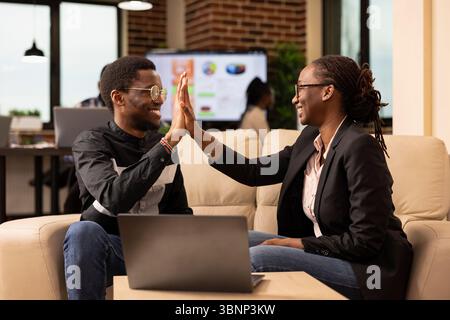I colleghi neri sorridenti si scambiano cinque alti, celebrando il successo del lavoro di squadra sul progetto aziendale. Allegro uomo d'affari e donna seduti sul divano, evidenziando una collaborazione efficace nell'ufficio di avvio. Foto Stock
