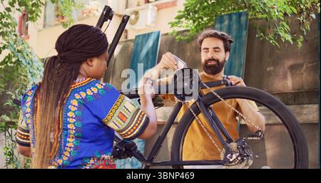 Ragazza afroamericana che assiste il fidanzato caucasico con una catena di biciclette arrugginita e lubrificante all'esterno come manutenzione estiva. Catena sportiva multietnica per l'ingrassaggio delle biciclette in cortile per una bicicletta sicura. Foto Stock