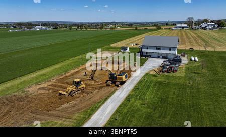 I macchinari pesanti operano in un cantiere vicino a una fattoria, con terreni in fase di preparazione per nuovi sviluppi sotto cieli luminosi e campi estesi Foto Stock