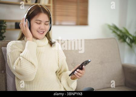 Giovane donna asiatica con cuffie che si rilassano a casa. ascolto di musica. seduti su un comodo divano con le cuffie Foto Stock