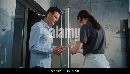 Lavoratore postale femminile che consegna piccoli pacchi per il destinatario in ufficio moderno, ottenendo la firma del cliente. Uomo d'affari che prende busta imbottita con ordine di acquisto online. Consegna o servizio postale. Foto Stock