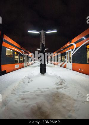 Moderni treni passeggeri arancioni sulla piattaforma ferroviaria innevata di notte, Estonia. Foto Stock
