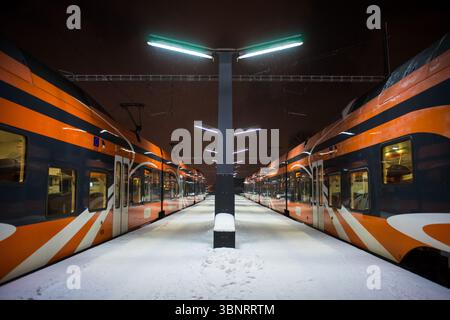 Moderni treni passeggeri arancioni sulla piattaforma ferroviaria innevata di notte, Estonia. Foto Stock