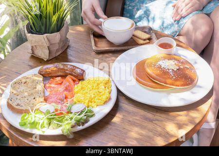 Una sostanziosa colazione americana mattutina è un piatto con uova strapazzate, salsiccia fritta e salmone affumicato su un tavolo di legno in un caffè all'aperto al sole Foto Stock