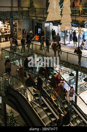 Londra, Regno Unito, 16 dicembre 2024. Gli amanti dello shopping fanno un giro sulle scale mobili in un affollato centro commerciale nel centro di Londra, visto dall'alto in bianco e nero. Foto Stock