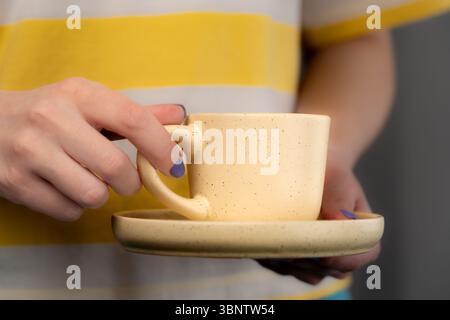 Una donna con una maglietta gialla e bianca con smalto viola regge una tazza di giallo pallido e un piattino con caffè. Primo piano. Foto Stock