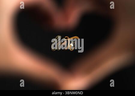 Amore. La parola "amore" brilla di lettere dorate, viste attraverso una forma a cuore fatta con le mani. Una fotografia romantica e creativa in primo piano. Foto Stock