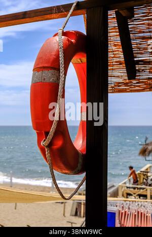 Salvagente rosso appeso su una capanna in legno con il mare e la spiaggia sabbiosa sullo sfondo. Giorno estivo luminoso con forte luce solare e atmosfera da spiaggia Foto Stock
