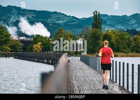Uomo in camicia rossa che fa jogging su un ponte panoramico in legno sul lago, con lussureggianti alberi verdi e colline sullo sfondo, vicino a Zurigo, Svizzera. Foto Stock