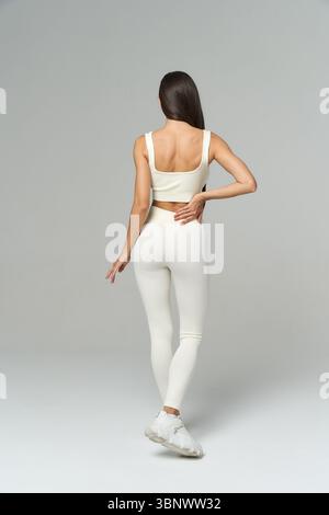 Donna in piedi con la schiena alla fotocamera, indossa un abbigliamento atletico bianco che include leggings e Crop top, in posa con una mano sull'anca. I capelli sono lunghi e scuri, Foto Stock