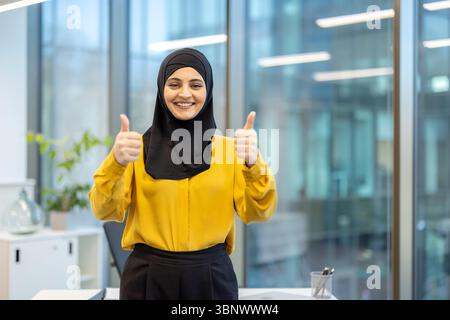 Una donna sorridente in un hijab dà un gesto con il pollice in alto in un ufficio. È felice e sembra sicura di sé. Foto Stock