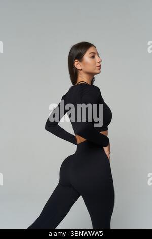 La donna in un abbigliamento sportivo nero aderente si pone con sicurezza in una vista laterale del profilo. La sua mano destra poggia sull'anca, evidenziando il suo fisico tonico. NEU Foto Stock