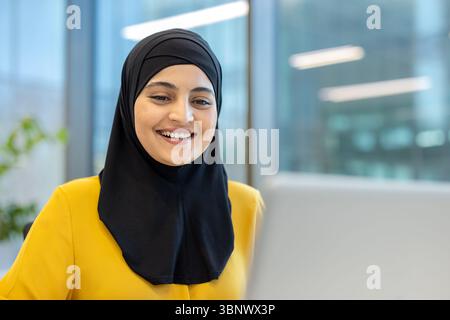 Una donna sorridente che indossa un hijab nero e una camicetta gialla guarda verso la fotocamera in interni. Foto Stock
