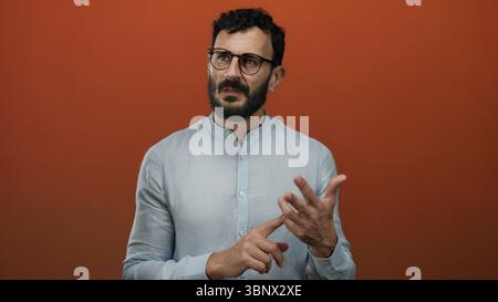Uomo con barba e occhiali in piedi su sfondo arancione, contando sulle dita, indossando camicia blu, ispanico, espressione premurosa, gesting, isol Foto Stock