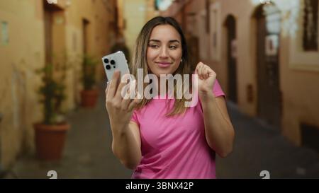 Una giovane donna che scatta un selfie con lo smartphone in una vivace strada all'aperto circondata da pareti dai colori caldi e piante in vaso, catturando un momento di gioia Foto Stock
