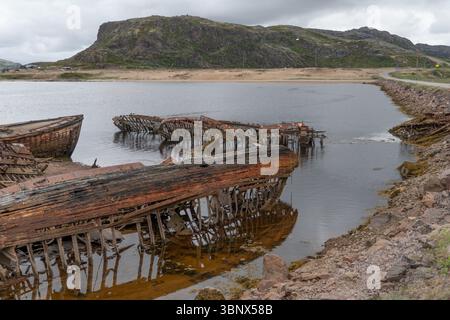 Relitti in legno arrugginito e metallo parzialmente sommersi in acque costiere poco profonde sullo sfondo di colline rocciose e cieli nuvolosi Foto Stock
