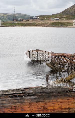 Relitti in legno arrugginito e metallo parzialmente sommersi in acque costiere poco profonde sullo sfondo di colline rocciose e cieli nuvolosi Foto Stock