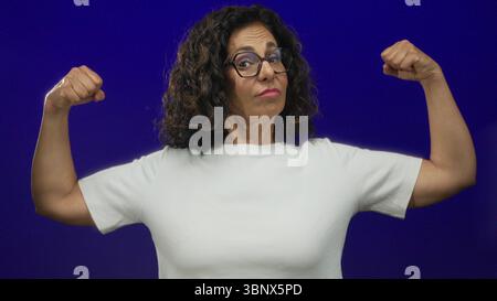 Donna ispanica di mezza età che flessa con sicurezza i muscoli contro un muro isolato di sfondo blu, mostrando forza e potenza nel suo casual Foto Stock