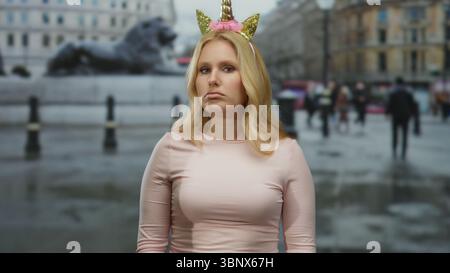 Una donna con diadema unicorno si trova in una strada cittadina che sembra divertente e preoccupata, sullo sfondo della vita urbana, trasmettendo un contrasto stravagante tra Foto Stock