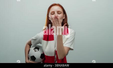 Una donna con i capelli rossi che regge una palla da calcio lancia un bacio con una sciarpa rossa e bianca, appoggiata su un semplice sfondo bianco. Foto Stock