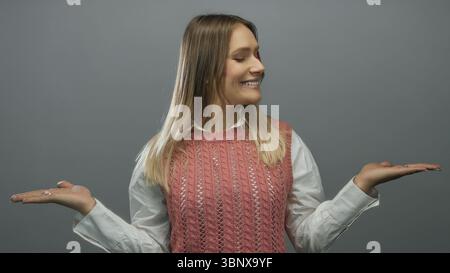 Donna sorridente che regge su sfondo grigio isolato indossando gilet rosa e camicia bianca che trasuda felicità e positività Foto Stock