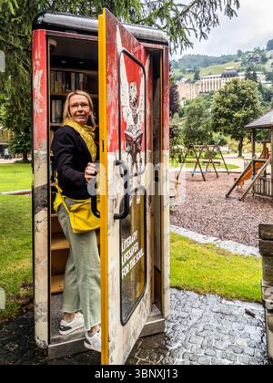 Un tempo una cabina telefonica, ora una stazione di scambio libri per lo scambio di libri presso i giardini termali di Engelberg. Una donna si trova in una cella pubblica di libri a Engelberg, in Svizzera, vicino al Titlis Centre, che funge da stazione di scambio libri. La cella dal design artistico ti invita a leggere, mentre sullo sfondo sono visibili un parco giochi e il paesaggio montano Foto Stock