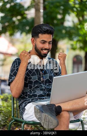 Giovane indiano che usa un computer portatile per celebrare la vittoria di buone notizie sui messaggi, la vittoria del jackpot della lotteria, giveaway online all'aperto. Il giovane turista felice si siede nel parco urbano della strada. Stili di vita della città. Foto Stock