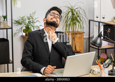 Uomo d'affari indiano libero professionista che si sente male malato malato affetto da mal di gola ferito deglutizione dolore tocca il collo lavorare a casa ufficio sul posto di lavoro. Maschio indù in tuta formale ha un raffreddore, influenza, virus Foto Stock