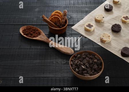 Primo piano dell'impasto per biscotti su una griglia da forno con cucchiaio di legno di cacao su un asciugamano da cucina, ciotola con gocce di cioccolato e misuratori in metallo vista frontale Foto Stock